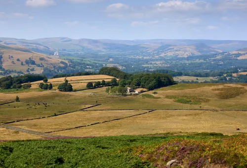 This landscape photograph captures Hope Valley in the Peak District, England, taken on a late summer morning. The image prominently features a farm in Hope Valley, with lush fields and stone walls stretching across the countryside. The Hope Cement Works factory is visible in the distance, serving as a recognizable landmark in the region. The natural beauty of the Peak District is highlighted by the blooming heather in the foreground, with rolling hills and clusters of trees. The scene is set within the context of the Longshaw Estate area, demonstrating the rural charm of the United Kingdom and its agricultural heritage.